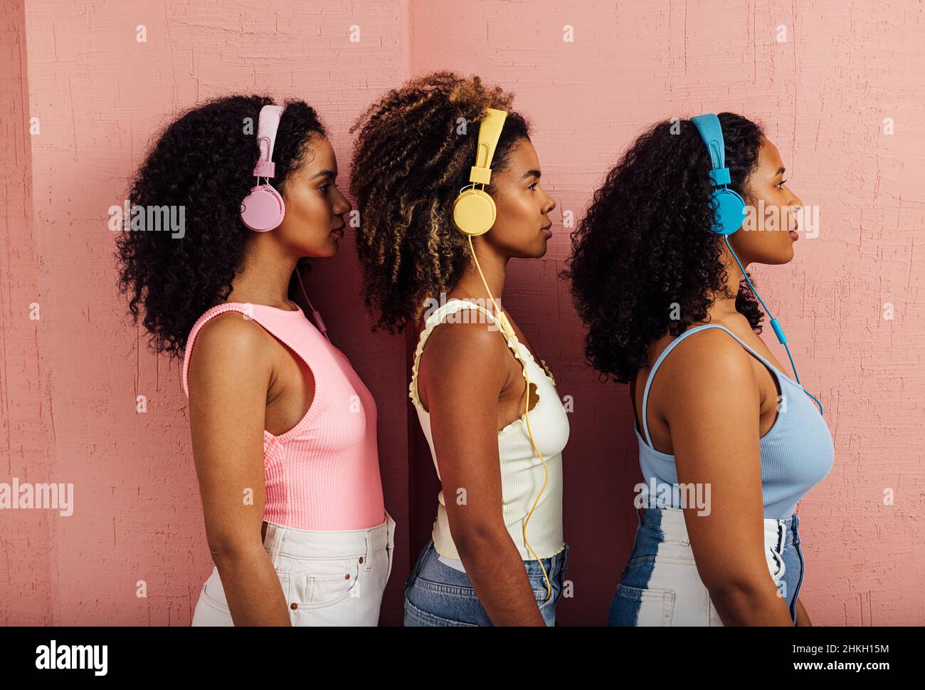Vista laterale di tre donne che indossano cuffie in piedi una dopo l'altra contro una parete rosa Foto Stock