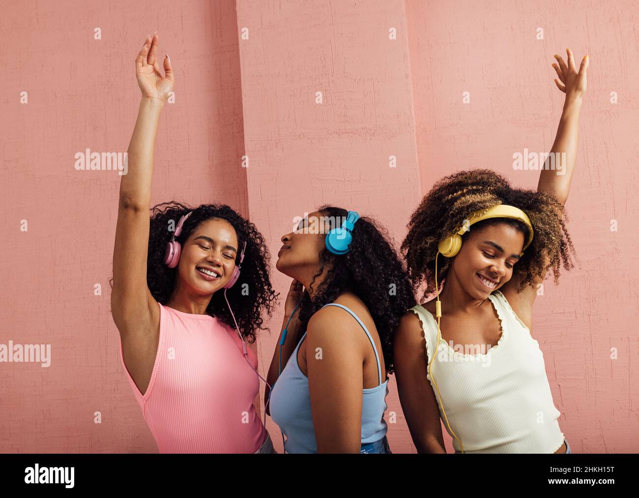 Gruppo di giovani donne che indossano cuffie e ballano insieme. Tre femmine in casuali che si godono la musica contro la parete rosa. Foto Stock