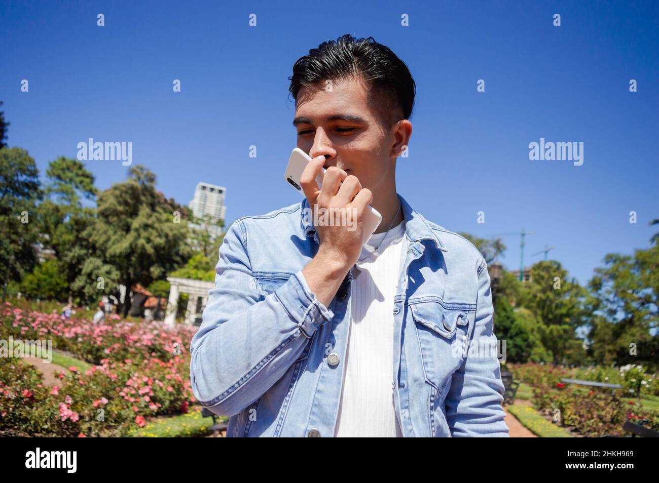 ritratto giovane argentino caucasico fuori a piedi in un parco con rose, fiori e alberi, innamorato dei suoi occhi chiusi baciando il telefono, felice Foto Stock