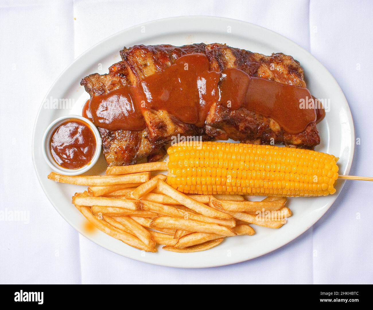 Costelinha de porco ao molho barbecue com batatatas fritas e milho em fundo de madeira branca. Foto Stock