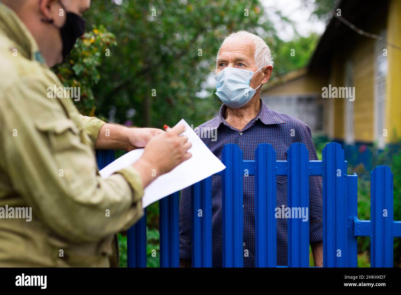 Uomo anziano in maschera protettiva comunicante con il rappresentante della compagnia di assicurazioni mentre si trova alla recinzione della sua casa di campagna Foto Stock