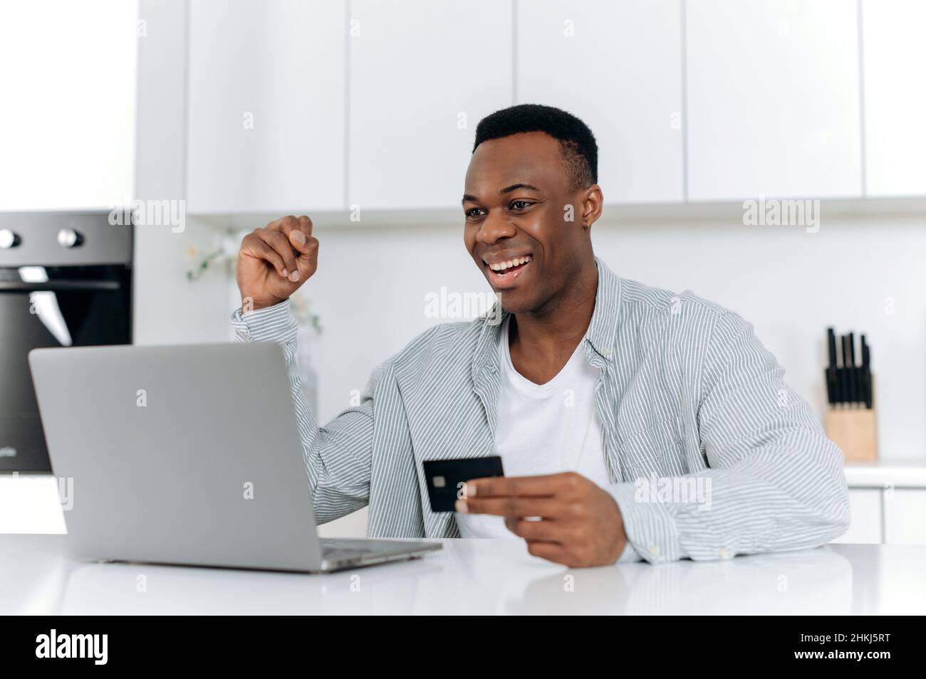 Shopping su Internet. Attraente, soddisfatto, giovane, adulto, ragazzo americano africano, seduti a un tavolo in cucina, facendo acquisti online su siti e applicazioni, inserendo i dettagli della carta, sorridendo. Foto Stock