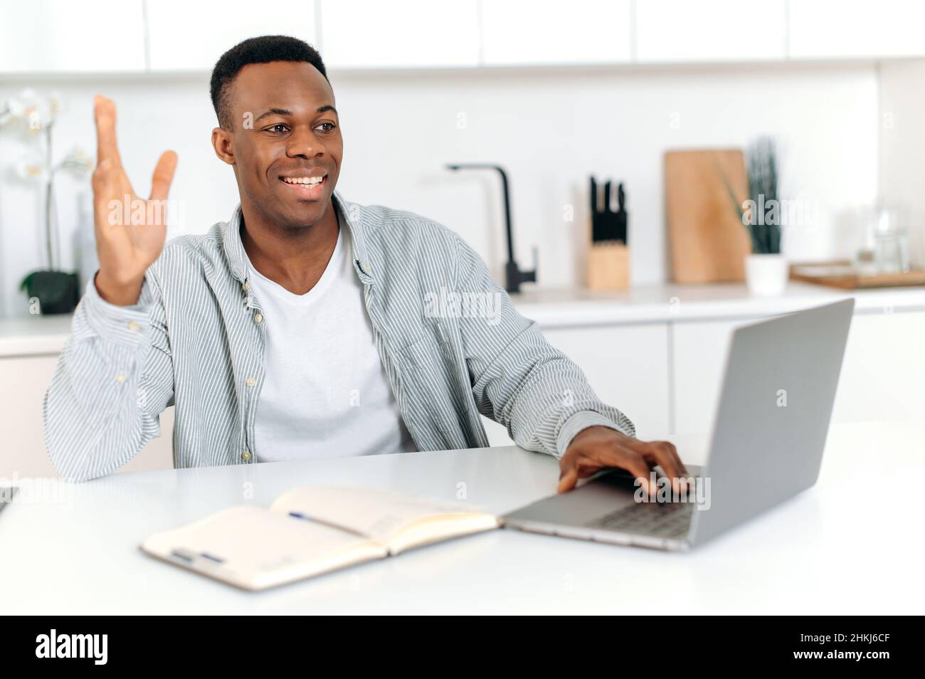 Il giovane afro americano di successo, freelance, designer o specialista IT, lavora da casa, utilizzando un computer portatile per comunicare con colleghi o clienti, sorridendo amichevole. Comunicazione a distanza Foto Stock