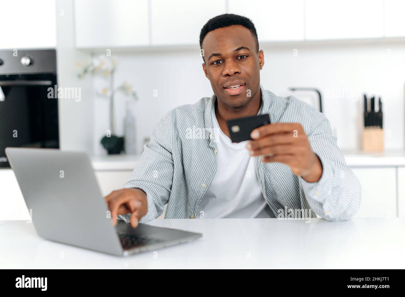 Positivo afroamericano giovane maschio adulto, seduto a un tavolo in cucina, utilizzando un computer e una carta di banca per pagare gli acquisti e le consegne online, inserendo dati, guardando una carta, sorridendo. Foto Stock