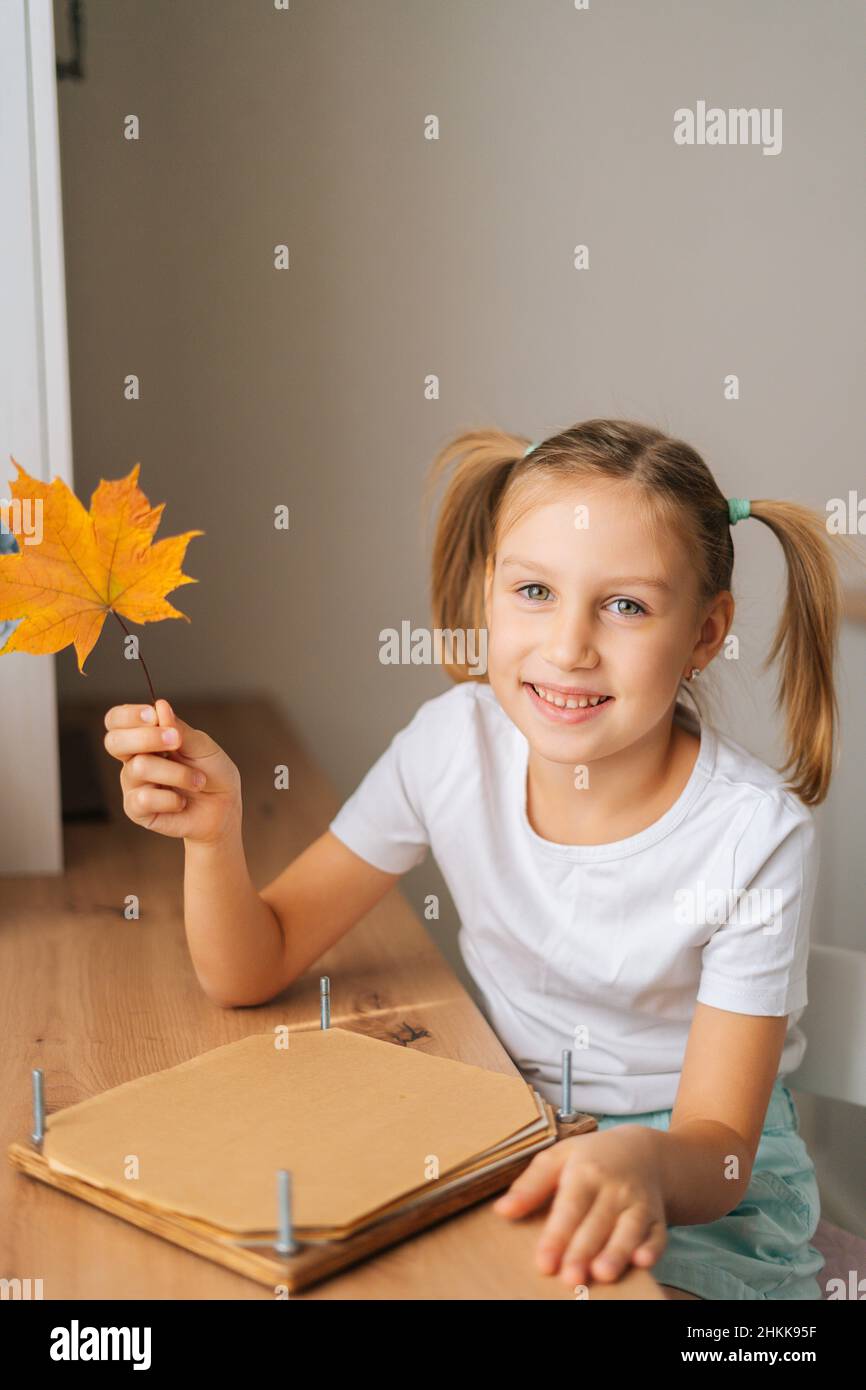 Ritratto verticale di sorridente bambina che tiene bella foglia di acero secco per creare erbario fai da te a casa. Foto Stock