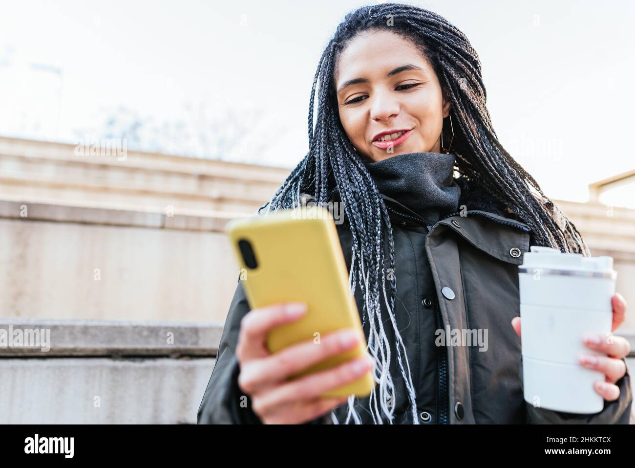 Positiva femmina ispanica con dreadlock che naviga in internet su un cellulare moderno mentre si siede su scale di legno con thermos bianchi in mano Foto Stock