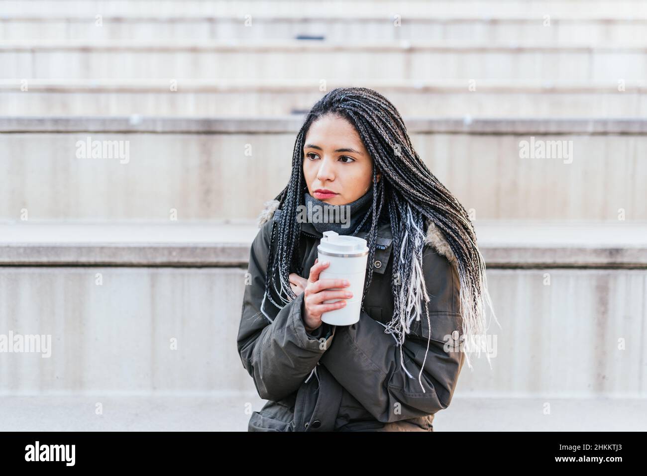 Donna ispanica in outerwear caldo con dreadlock guardare via mentre si siede sulle scale con thermos di caffè caldo in città in tempo freddo Foto Stock