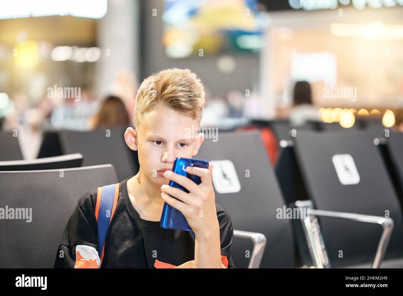 Ragazzo adolescente con acconciatura bionda corta e zaino frena con espressione facciale seria e legge ebook via telefono in aeroporto Foto Stock