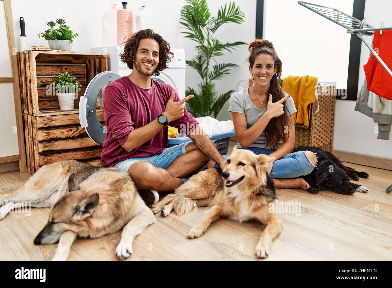 Giovane coppia ispanica che fa la lavanderia con i cani allegri con un sorriso sulla faccia che punta con la mano e il dito fino al lato con esposizioni felici e naturali Foto Stock