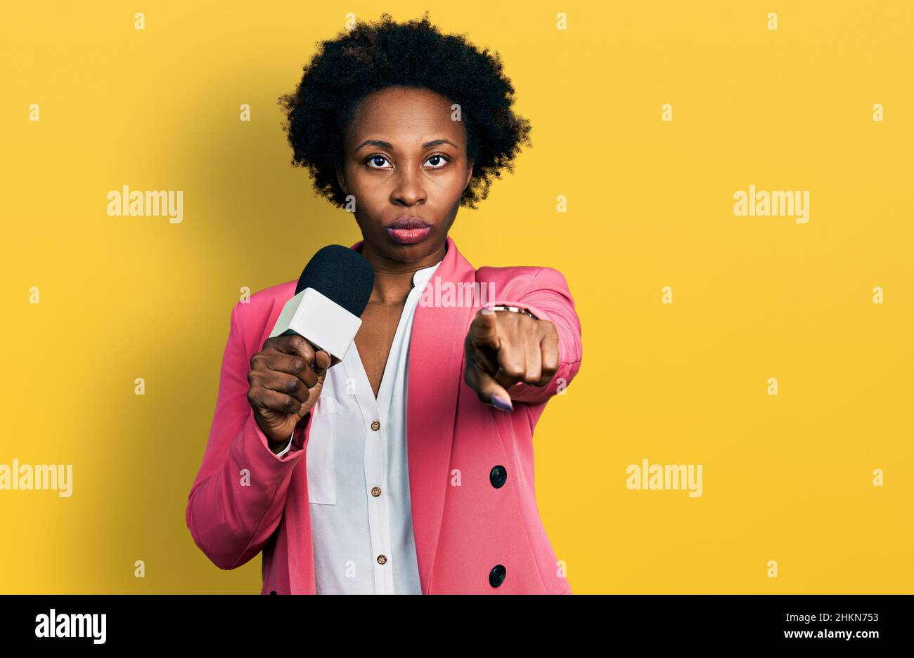 Donna afroamericana con capelli afrofroche tengono il microfono reporter che punta con il dito alla fotocamera e a voi, gesto sicuro guardando serio Foto Stock