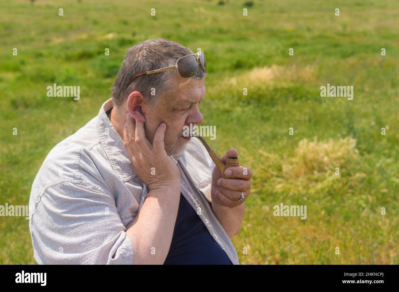 Ritratto con vista laterale del tubo del tabacco caucasico senior fumante mentre si siede all'aperto contro pascolo primaverile Foto Stock