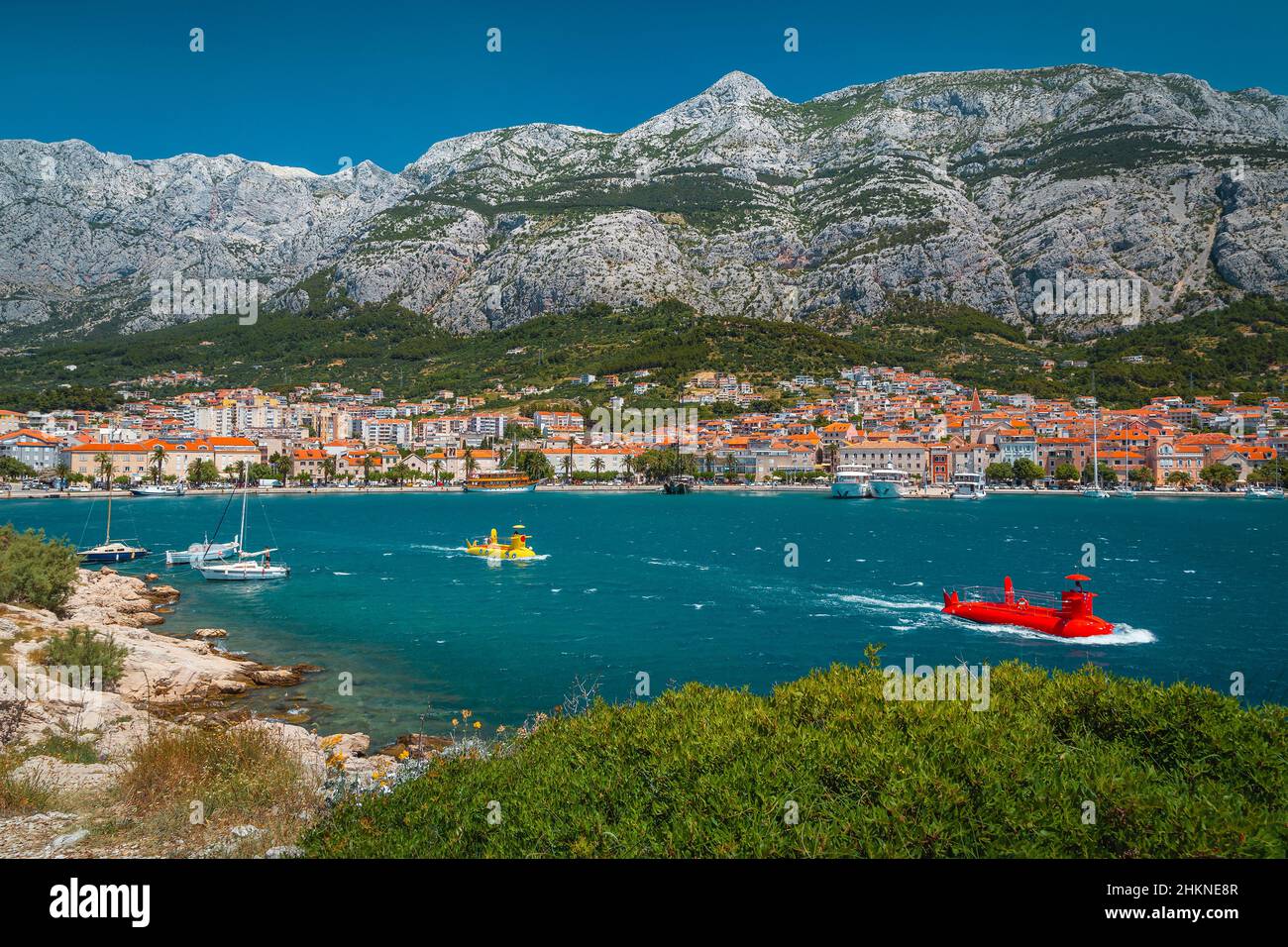 Variopinte imbarcazioni sottomarine che trasportano turisti vicino alla costa. Bella città di Makarska con porticciolo e alte montagne sullo sfondo, Dalmat Foto Stock