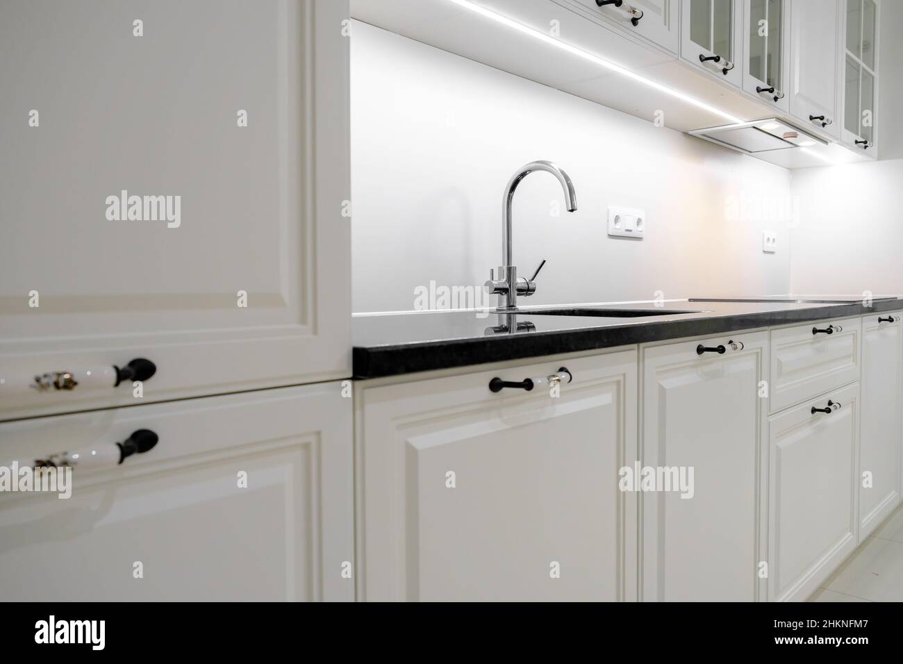 Vista laterale moderna ed elegante cucina con cassetti e armadi bianchi, piano di lavoro in marmo nero e lavello costruito con rubinetto d'argento Foto Stock