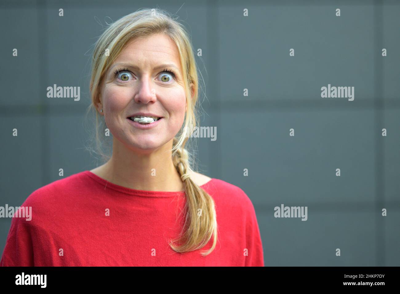 Donna bionda che si diverte con bubblegum o gomma da masticare con occhi larghi contro una parete grigia Foto Stock