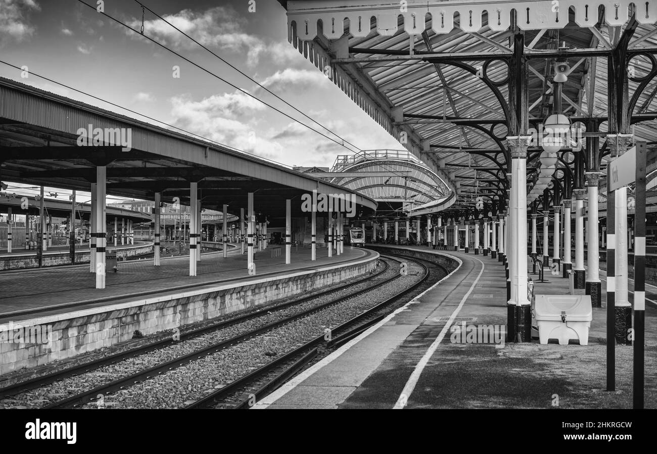 Una piattaforma storica della stazione che si curva verso un treno d'attesa. C'è un nome di luogo in primo piano e cielo con le nuvole sopra. Foto Stock