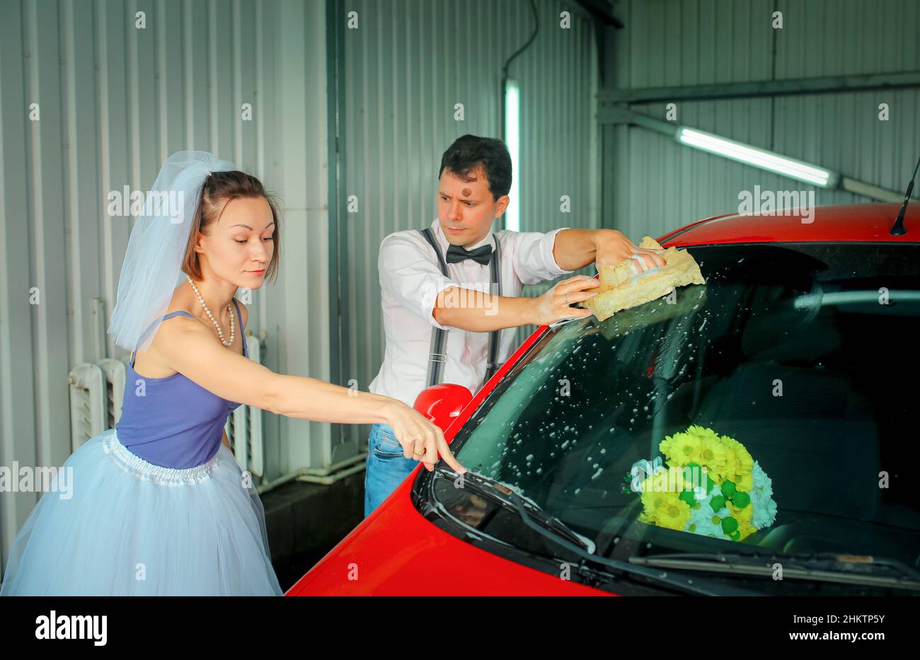 Un giovane uomo con una cravatta ad arco e una donna in un velo stanno lavando un'auto ad un autolavaggio. Anniversario di matrimonio. Umorismo. Foto Stock