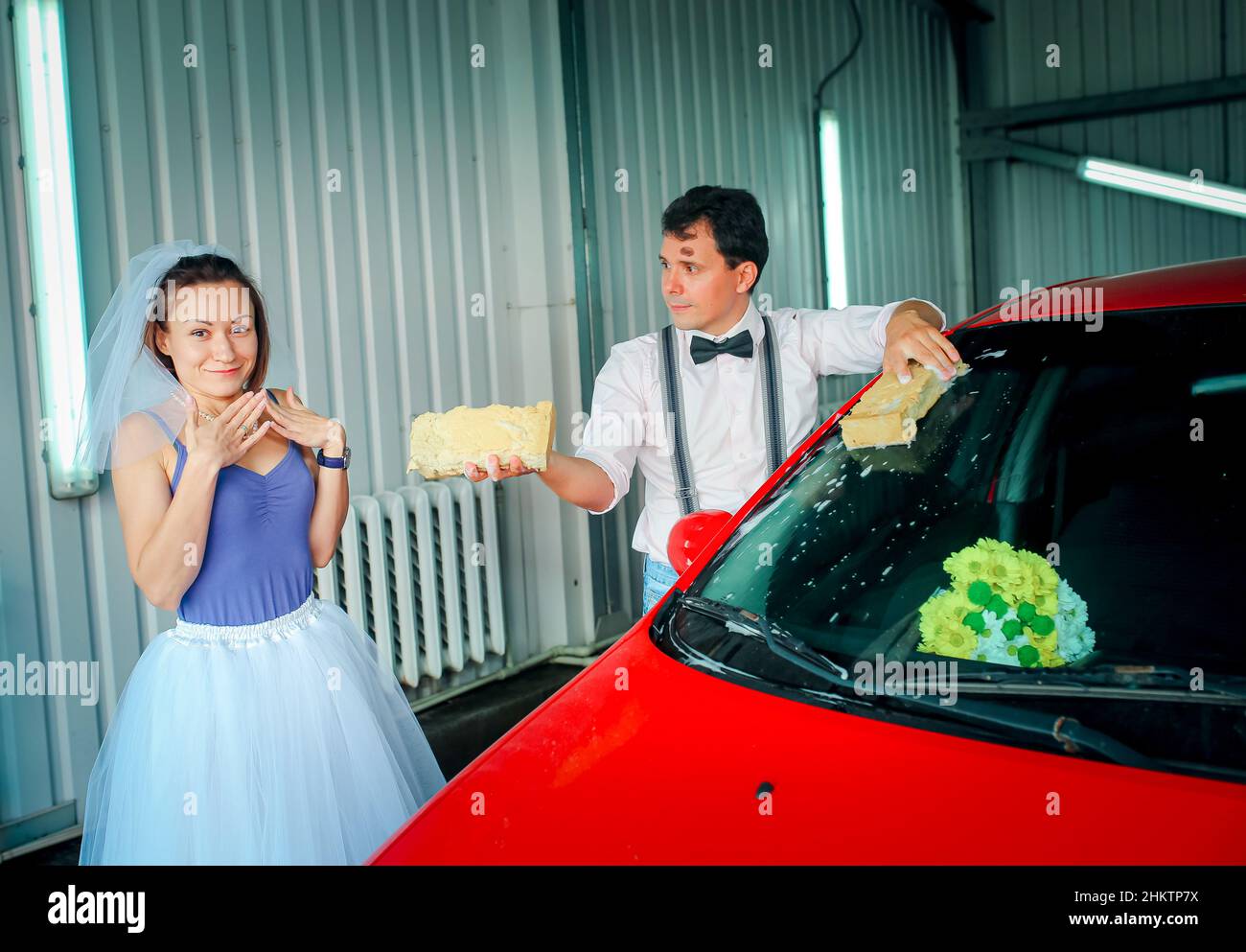 Un giovane uomo con una cravatta ad arco e una donna in un velo stanno lavando un'auto ad un autolavaggio. Anniversario di matrimonio. Umorismo. Foto Stock