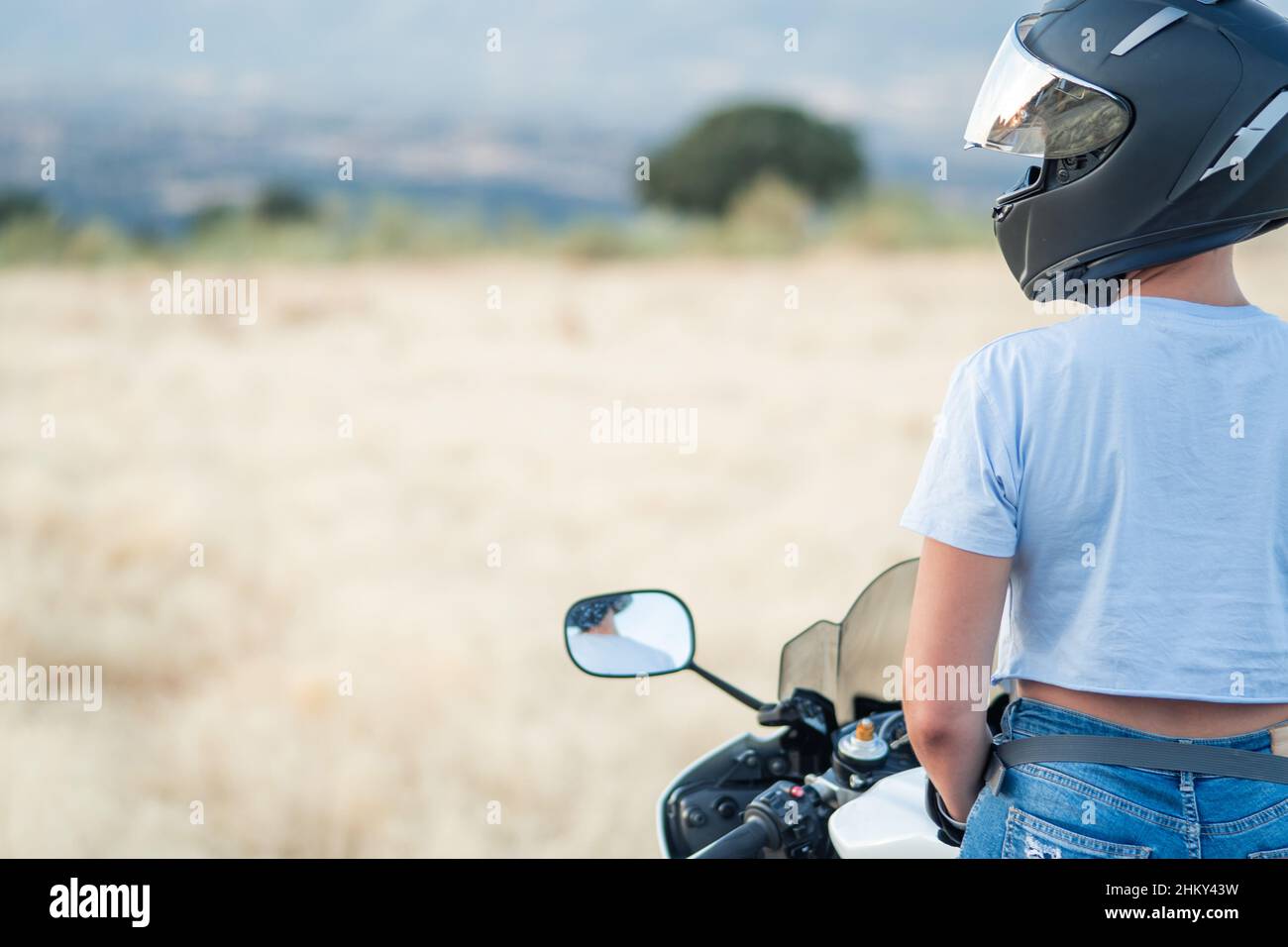 giovane donna biker da dietro con t-shirt blu e casco di sicurezza seduto sulla sua moto guardando l'orizzonte. spazio copia Foto Stock