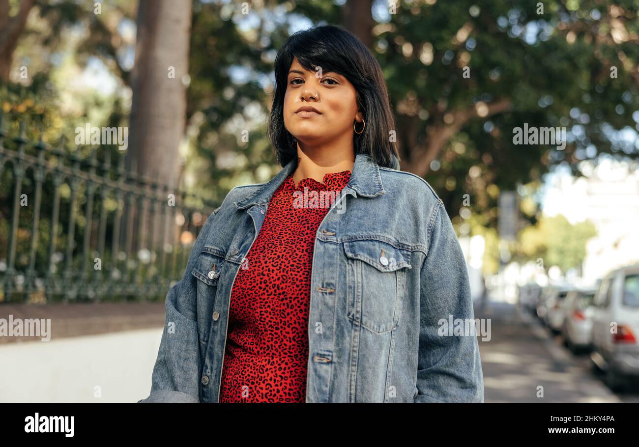 Donna sicura che guarda la fotocamera mentre si alza su un marciapiede in città. Donna di metà età adulta in piedi da sola accanto a un parco all'aperto. Donna Wea Foto Stock