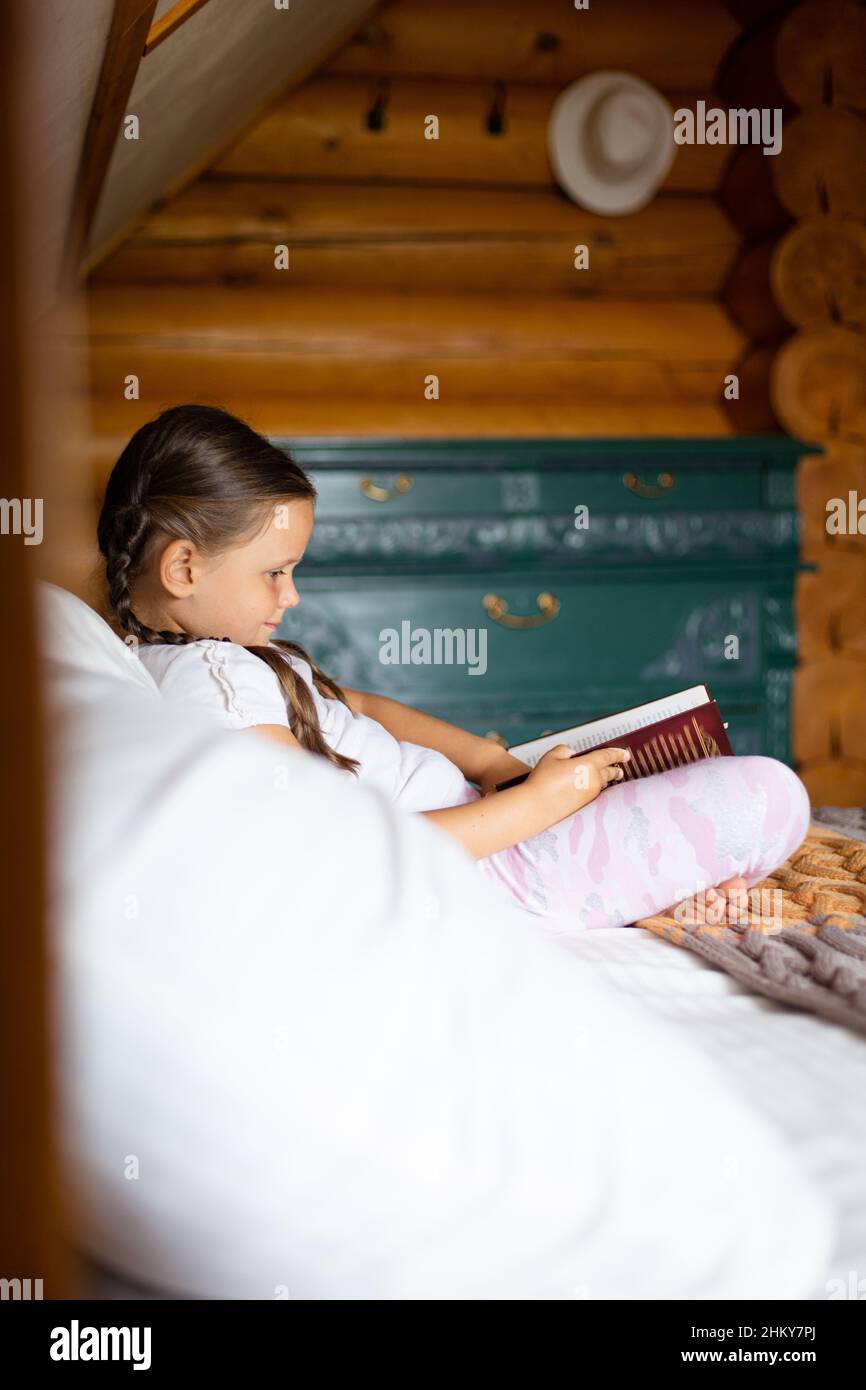 Ragazza giovane rilassata che non sa che è sulla macchina fotografica seduta sul letto doppio grande che tiene il libro nelle mani che guardano il libro che legge che porta i pigiami in legno Foto Stock