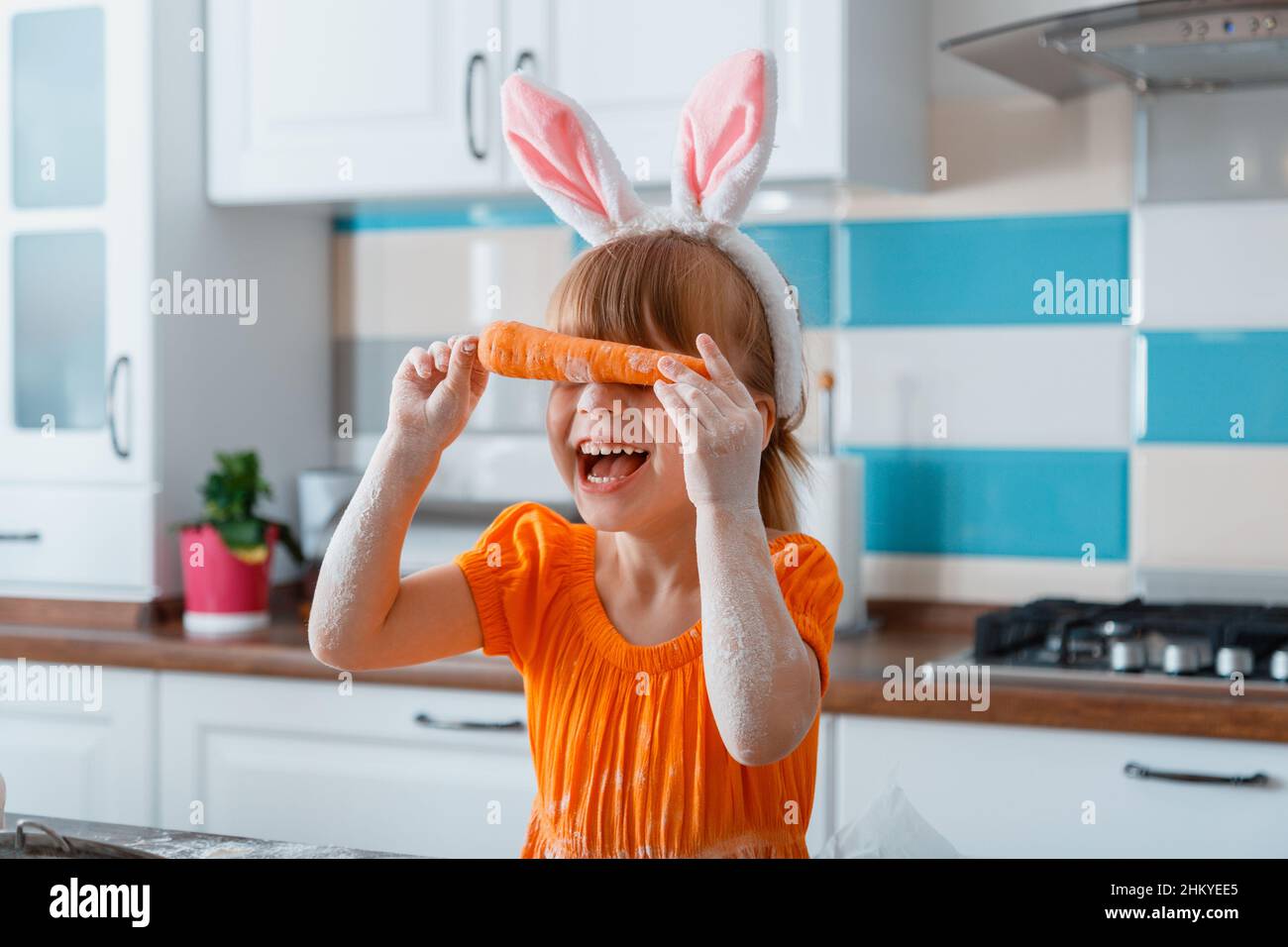 Ragazza bambino ride fare faccia divertente usando carota e coniglietto rosa orecchie. Ragazza figlia bambino ottenere sporco in farina in cucina mentre cucinando prima di Pasqua Foto Stock