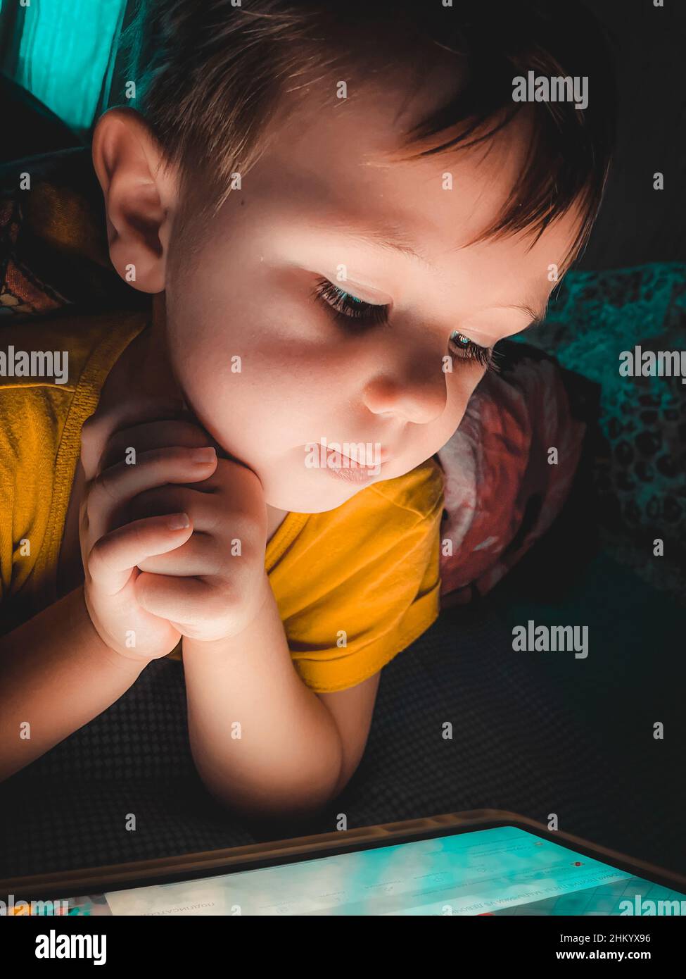 Piccolo ragazzo caucasico che guarda un tablet la sera al buio sul letto, fotografia stile di vita a casa, fuoco selettivo e softfocus Foto Stock