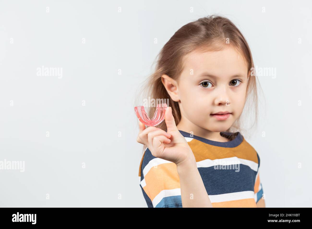 Carino ragazza caucasica piccola con i capelli biondi sta tenendo un addestratore dentario di miofunctional rosa sul bianco lo sfondo. Tariner dentale è fatto per aiutare equala Foto Stock