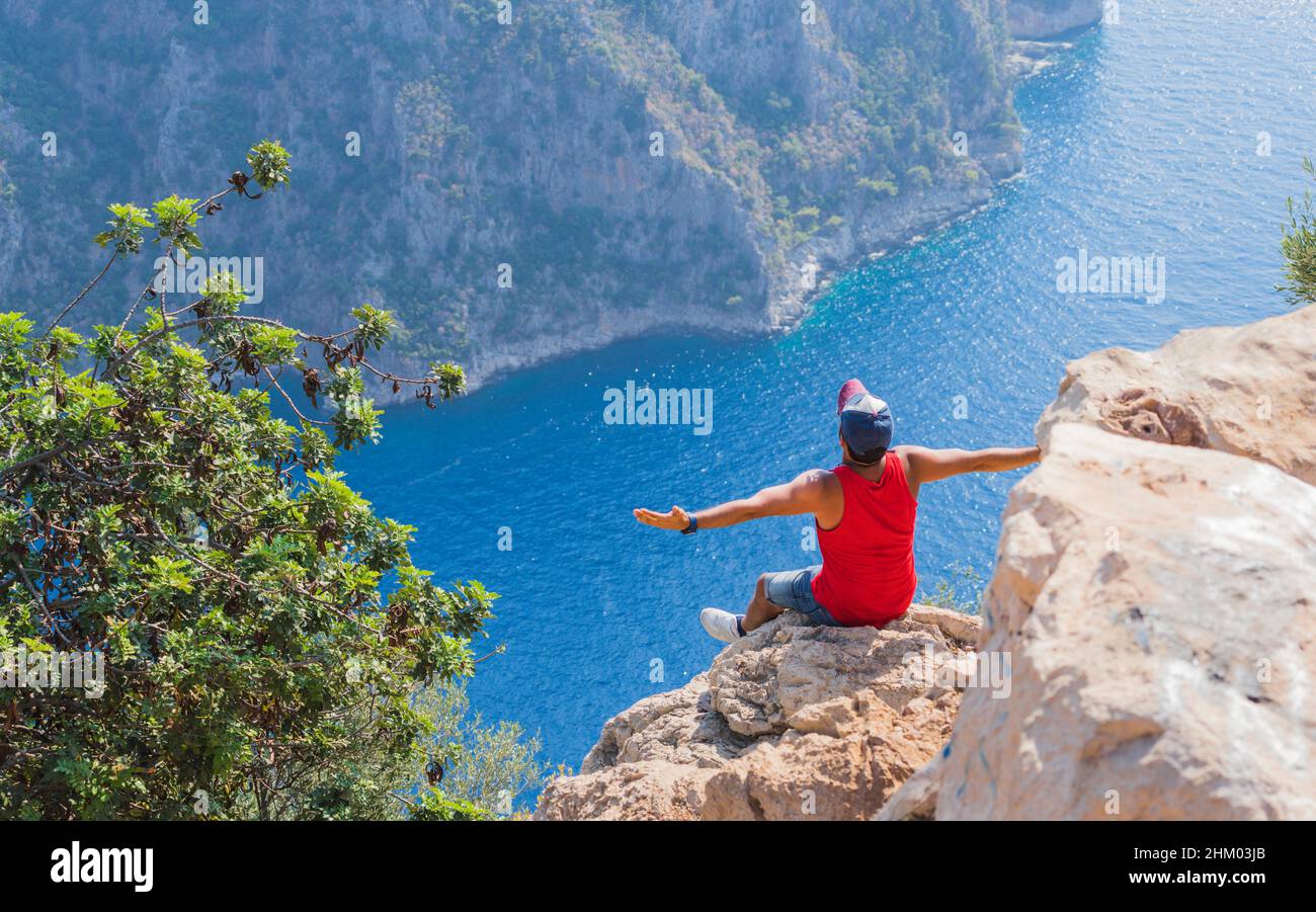 Giovane uomo seduto al bordo di una scogliera a braccia aperte e godendo il bellissimo paesaggio marino. Vista della Valle delle Farfalle in Turchia. Concetto di libertà. Foto Stock