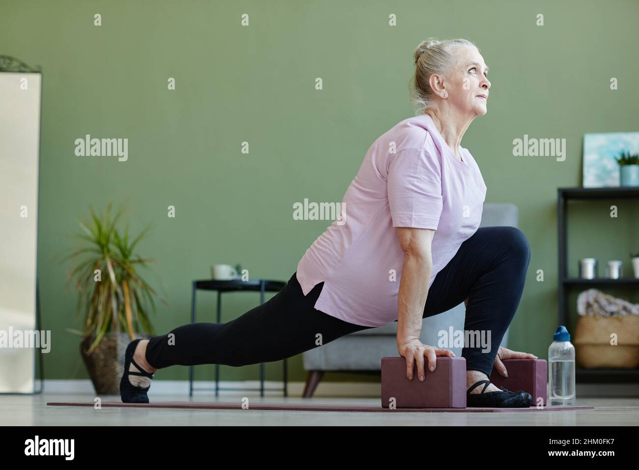 Donna anziana attiva con blocchi di yoga in piedi con una gamba stirata e un'altra piegata nel ginocchio mentre si esercita sul pavimento Foto Stock