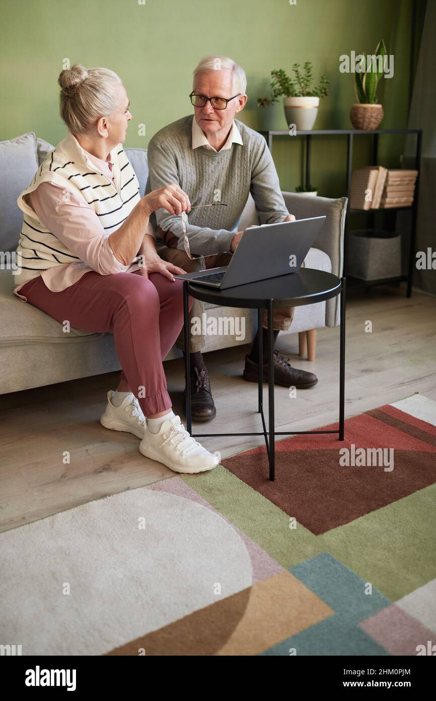 Due adulti anziani in abiti casual seduti sul divano di fronte al computer portatile in soggiorno e discutere di cosa acquistare nel negozio online Foto Stock