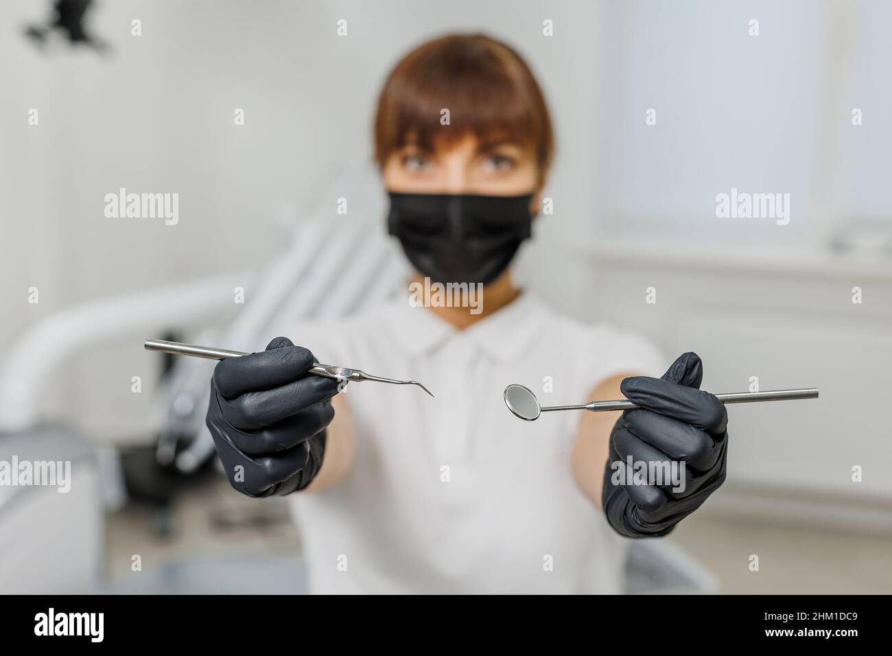 Donna dentista in uniforme bianca, maschera e guanti neri che mostrano gli strumenti di stomatologia tra le moderne apparecchiature dentarie. Ritratto di medico femminile - stomatologo professionista, ortodontista. Foto Stock