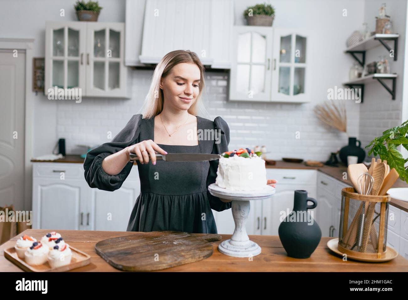 Pasticciere dello chef giovane donna caucasica in abito grigio con fetta di torta tagliata a coltello sul tavolo da cucina. Tortini e dolci dessert in stile scandinavo Foto Stock