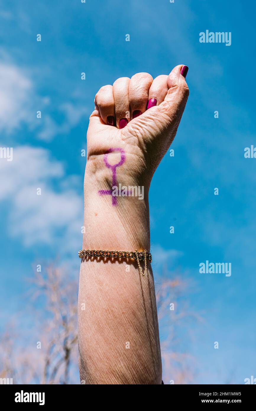 Pugno di una donna più anziana con chiodi dipinti di viola, con il cielo sullo sfondo, con il simbolo femminile dipinto. Concetto di giornata delle donne, empowerment, Foto Stock