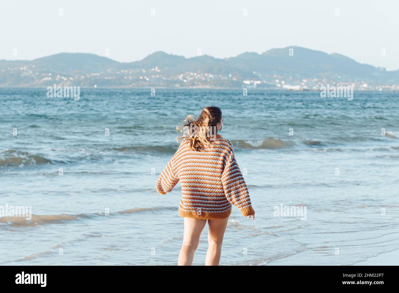 Una giovane donna sulla spiaggia con una maschera chirurgica durante una giornata intensa indossando un pullover con spazio per la copia e felice attitudine Foto Stock