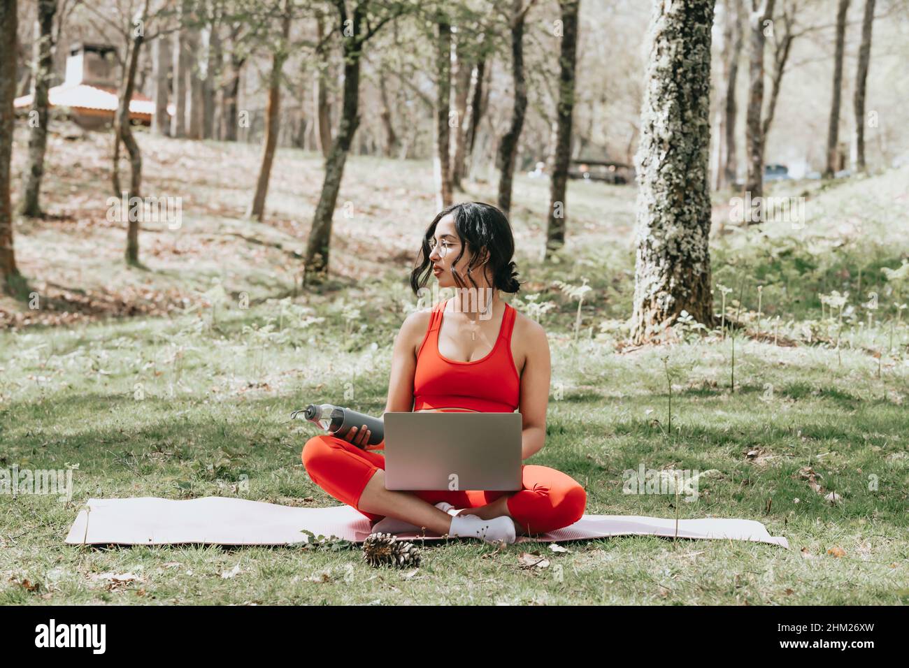 Giovane donna in abiti fitness rilassarsi sul suo computer portatile afferrare una bottiglia d'acqua al parco su un tappetino yoga Foto Stock