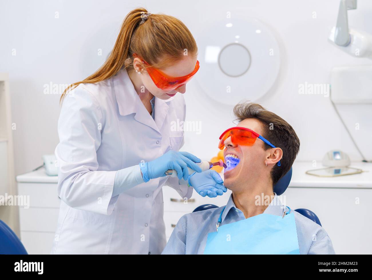 Medico femminile in uniforme medica con guanti in lattice e occhiali protettivi che mettono luce curante nella bocca aperta del giovane durante il lavoro in clinica dentale moderna Foto Stock