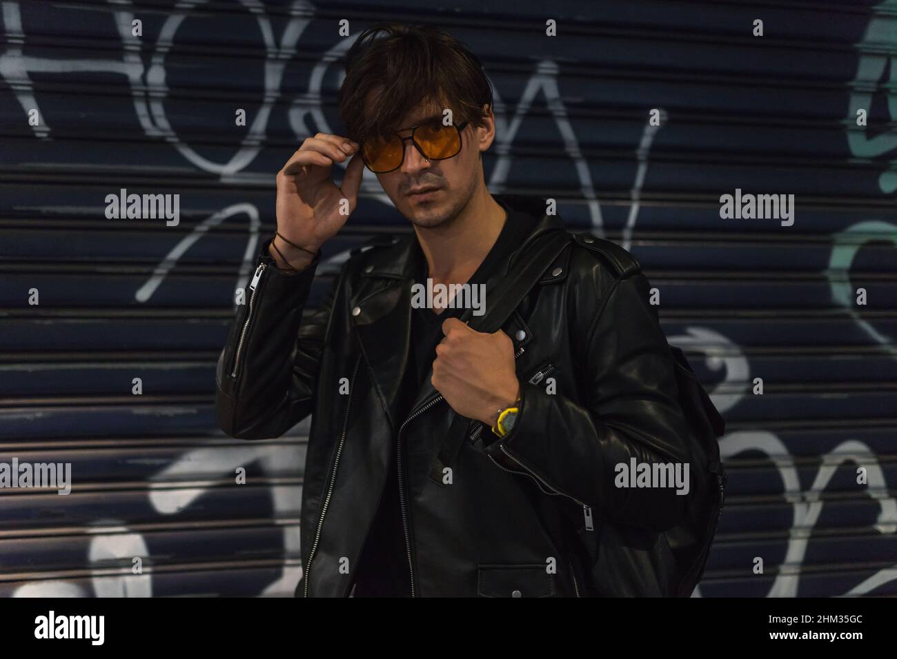 Ritratto di giovane uomo caucasico alla moda in giacca nera in pelle e occhiali da sole gialli. In piedi vicino al muro con graffiti. Foto di alta qualità Foto Stock