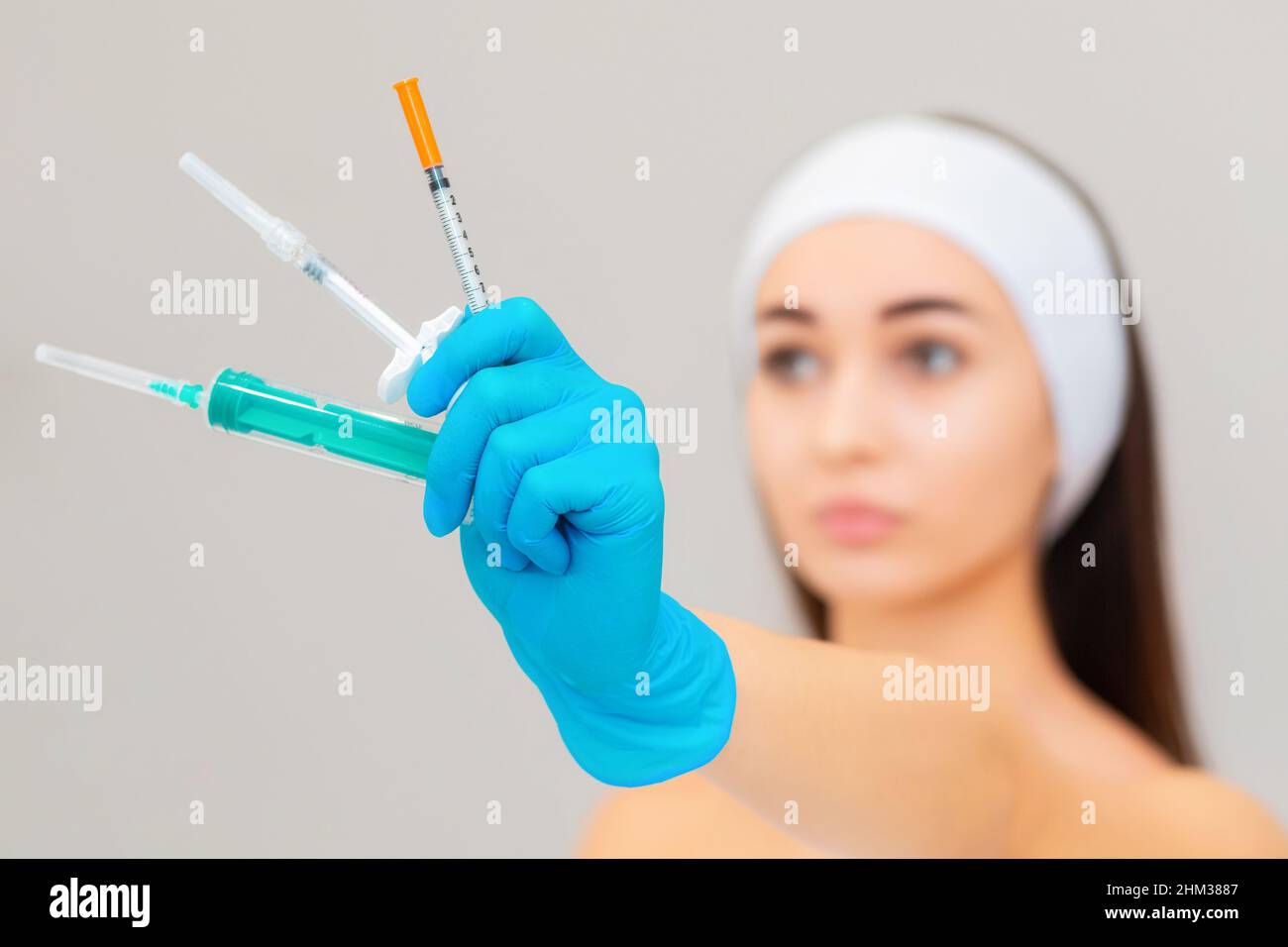 Un ritratto sfocato di una giovane donna caucasica con una mano guantata che tiene tre diverse siringhe. Sfondo grigio. Il concetto di bellezza e ringiovanimento Foto Stock