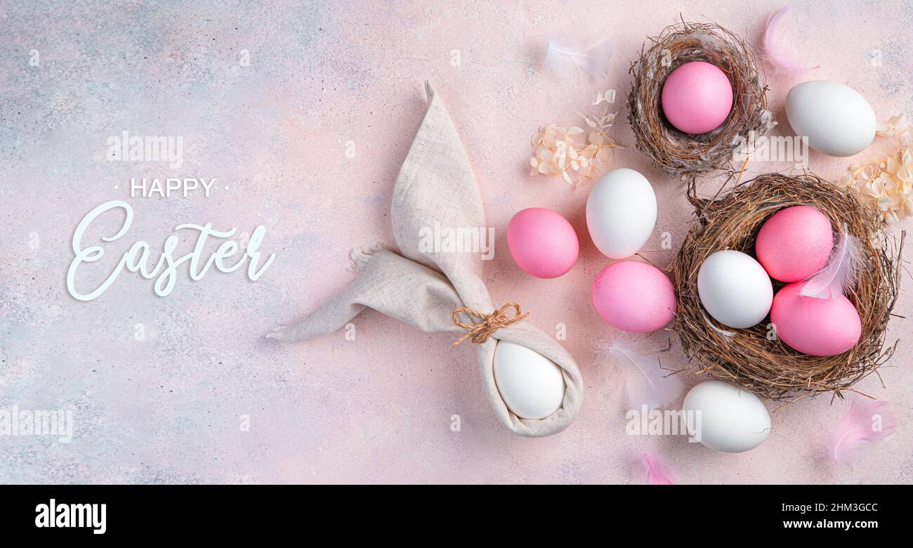 Buona Pasqua, uova, nido e coniglietto di Pasqua fatto di tovaglioli su sfondo rosa chiaro. Vista dall'alto. Foto Stock