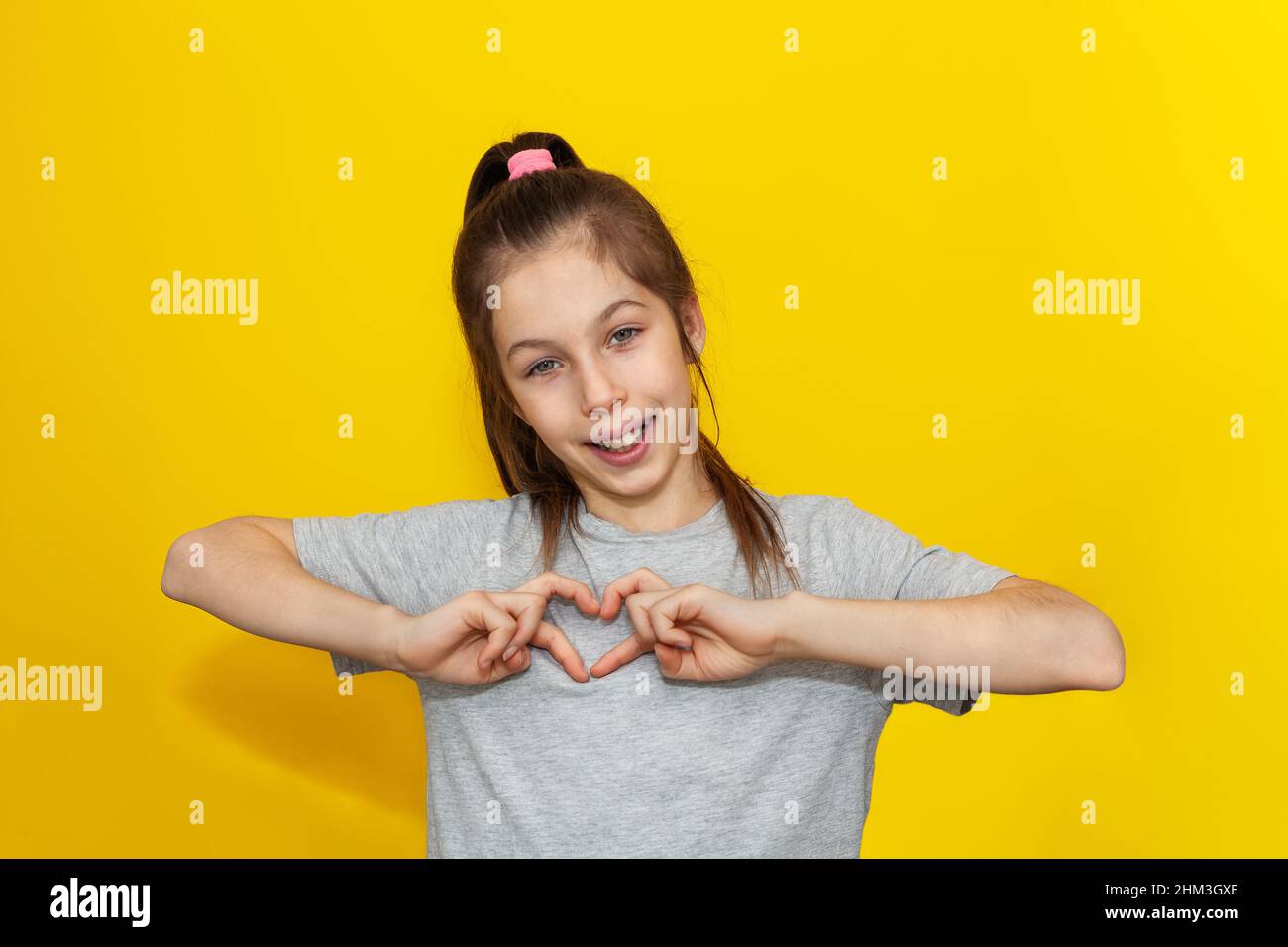 Adorabile ragazza in t-shirt grigia facendo un gesto di cuore con le dita con un sorriso felice e sincero isolato su sfondo giallo. Espressione dell'amore a Foto Stock