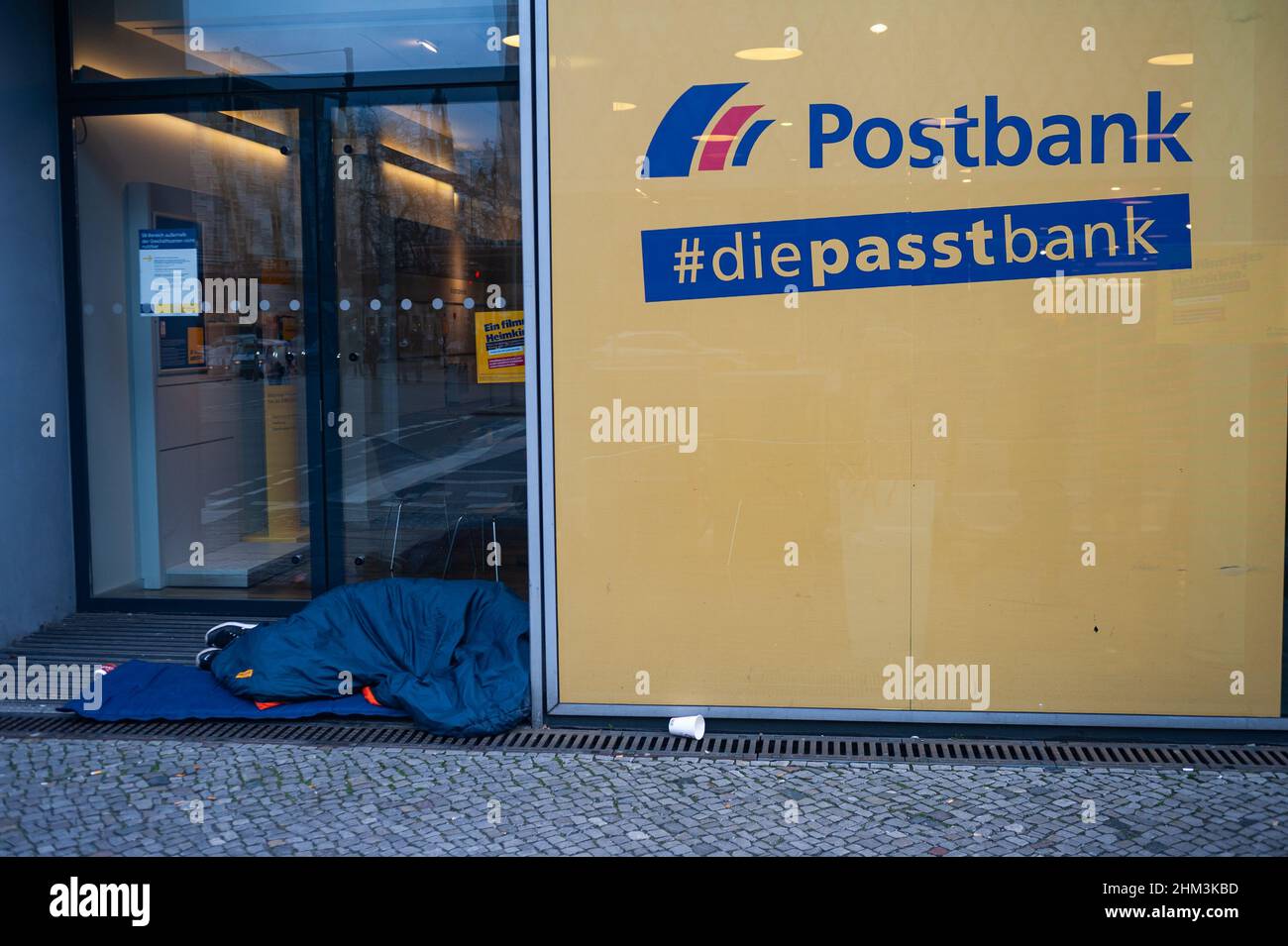 05.02.2022, Berlino, Germania, Europa - il senzatetto dorme nel sacco a pelo sul marciapiede di fronte a una filiale Postbank nel centro della città. Foto Stock