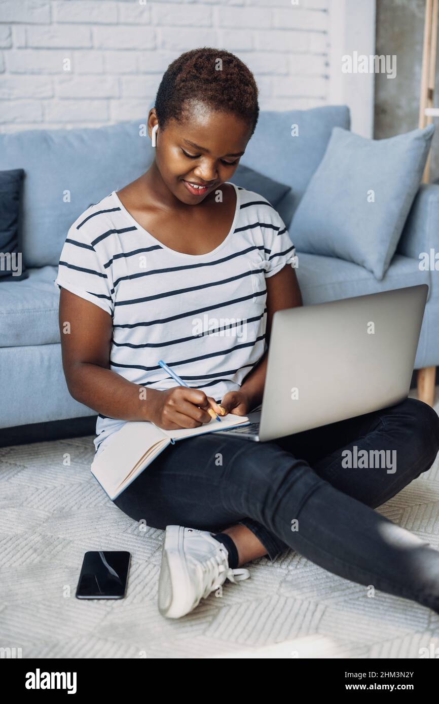 Donna afro che studia in remoto partecipando a una conferenza online e prendendo appunti in un notebook. Seduta sul pavimento utilizzando il suo computer portatile. Tecnologia Internet Foto Stock