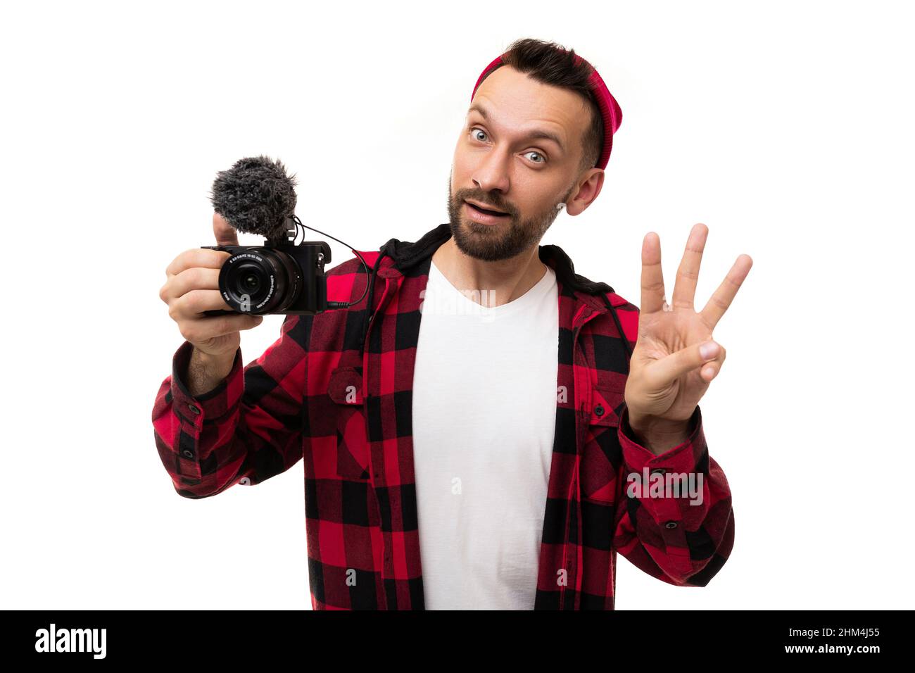 un blogger bearded uomo in un cappello rosso e camicia in una gabbia rossa nera tiene una macchina fotografica con un microfono nelle sue mani e mostra il numero tre con un Foto Stock