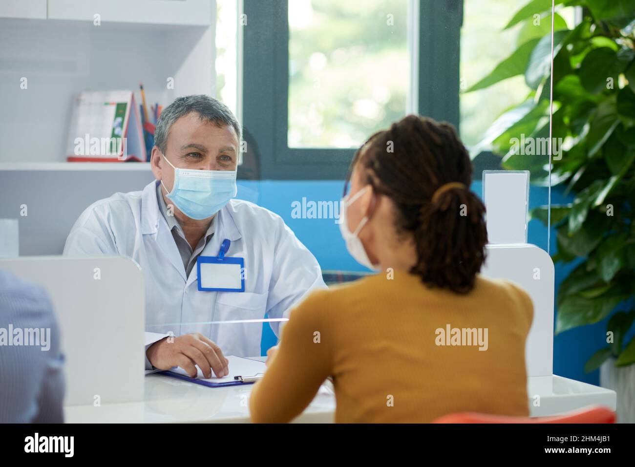Medico generico senior che fornisce raccomandazioni al paziente quando sono seduti con schermo di vetro tra loro a causa di coronavirus pandemia Foto Stock