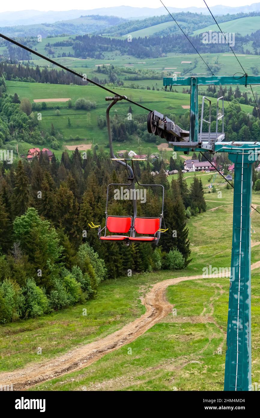 Seggiovia in una stazione sciistica sullo sfondo del paese e delle montagne. Ascensore nella stagione estiva. Corda e sedie in movimento. Pineta verde Foto Stock