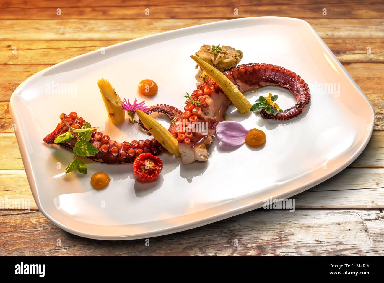 Tentacoli di polpo arrostiti con mais giovane, pomodoro e foglie di menta in piatto bianco su tavole di legno Foto Stock