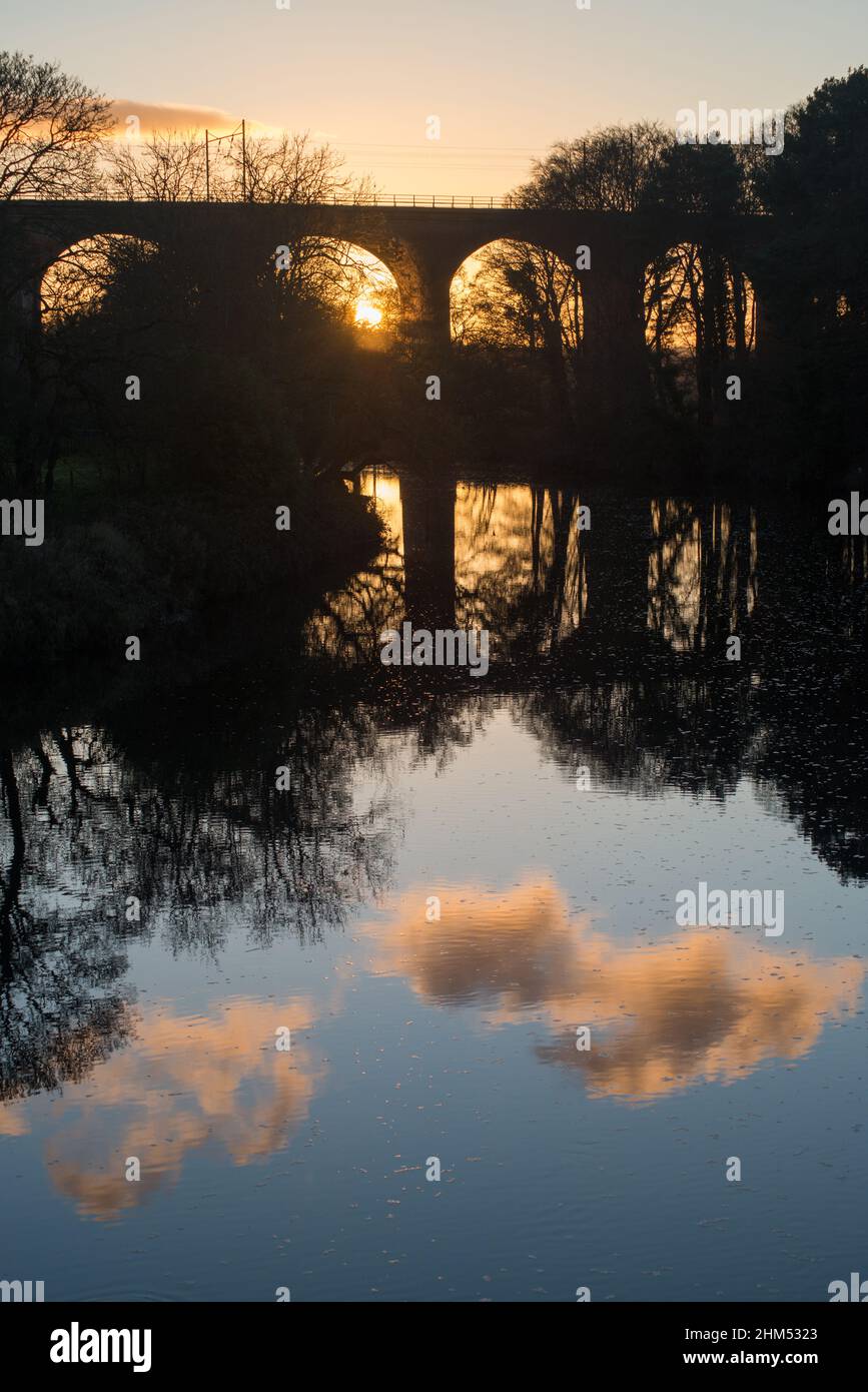 Immagine ritratto del tramonto visto attraverso archi di un viadotto con la struttura alberi e le nuvole dorate in silhouette e riflessi del fiume Foto Stock