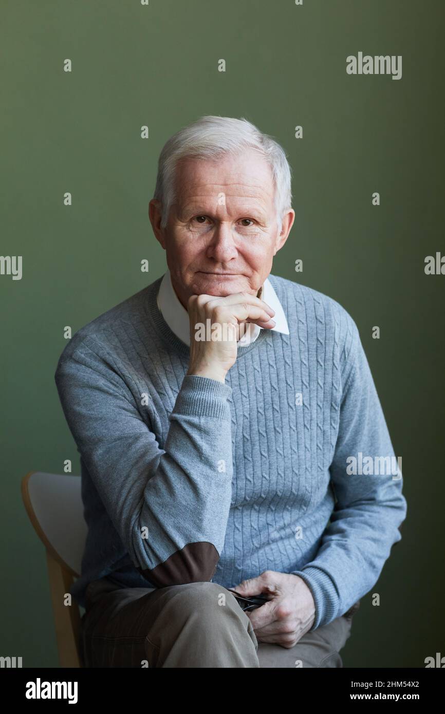 Uomo anziano serio in pullover azzurro seduto sulla sedia davanti alla macchina fotografica contro il muro verde scuro e tenendo la mano dal mento Foto Stock