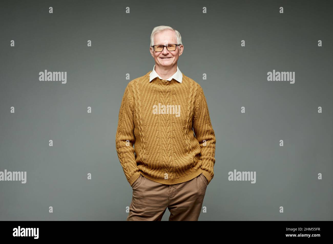 Uomo anziano felice in maglia maglione e pantaloni marroni in piedi su sfondo grigio e guardando la macchina fotografica con sorriso Foto Stock