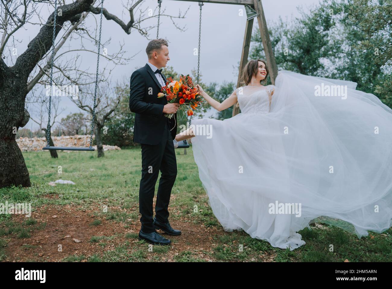 Felice coppia sorridente di stile che cammina in Toscana, Italia il giorno del matrimonio. SPOSI NOVELLI CON L'ALTALENA NEL PARCO. La sposa e lo sposo camminano giù il Foto Stock